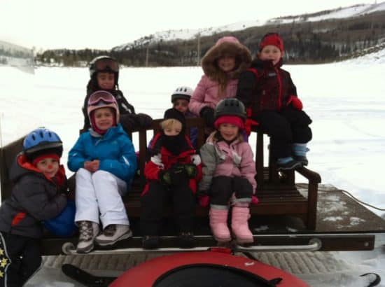 8 Kids sit on a sleigh being pulled by a snowmobile, heading out to go downhill tubing!