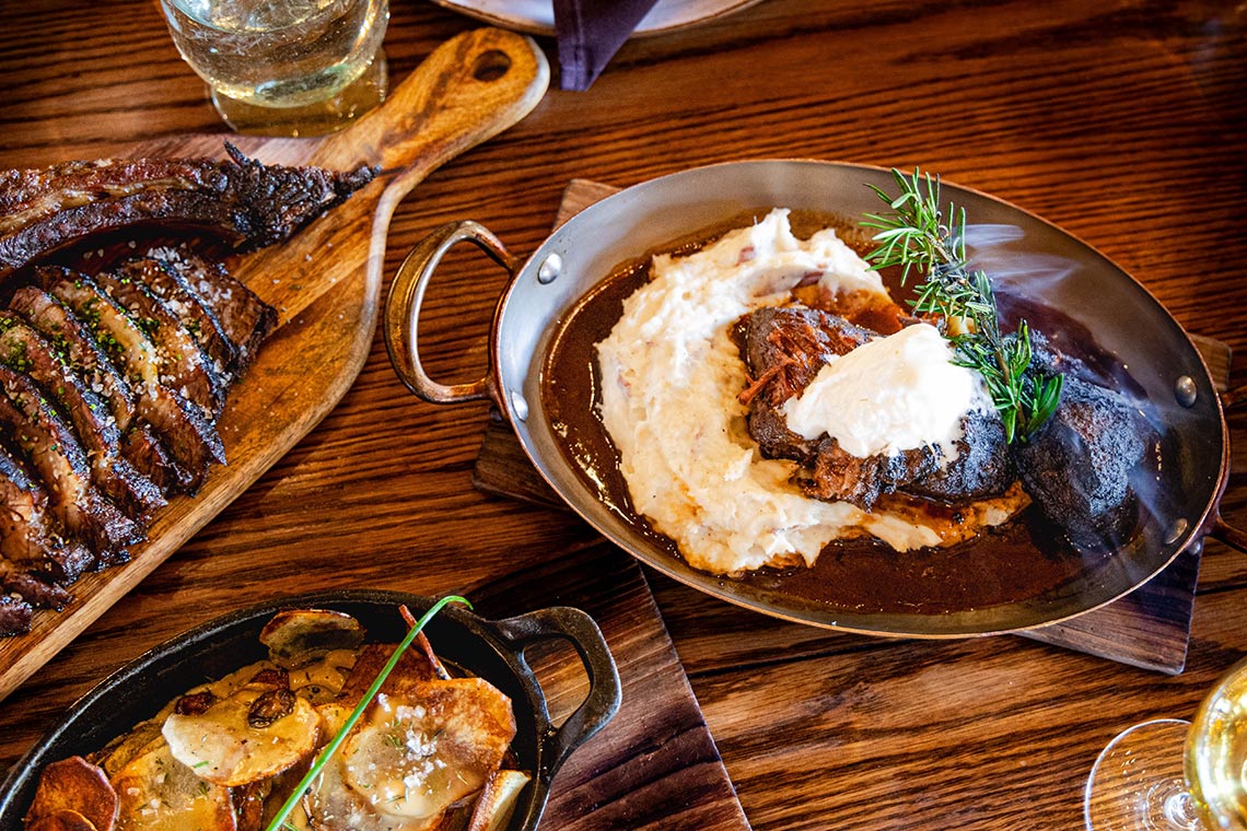Sliced stead on a wooden carving board, a dish of scalloped potatoes and another bowl of mashed potatoes and gravy.