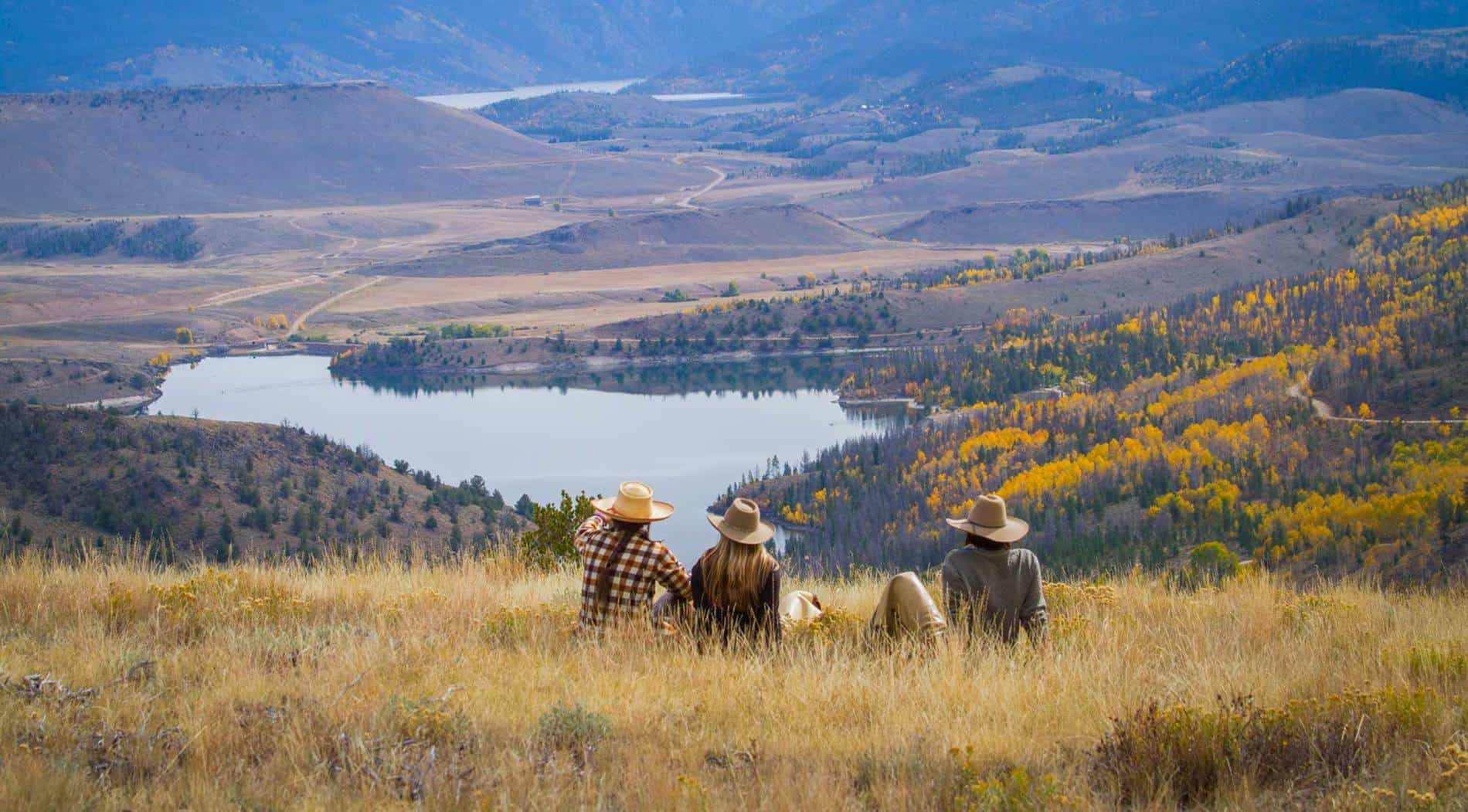 People with cowboy hats sit on a hillside that overlooks yellow aspen hillsides, a lake, and more of Grand county in the distance