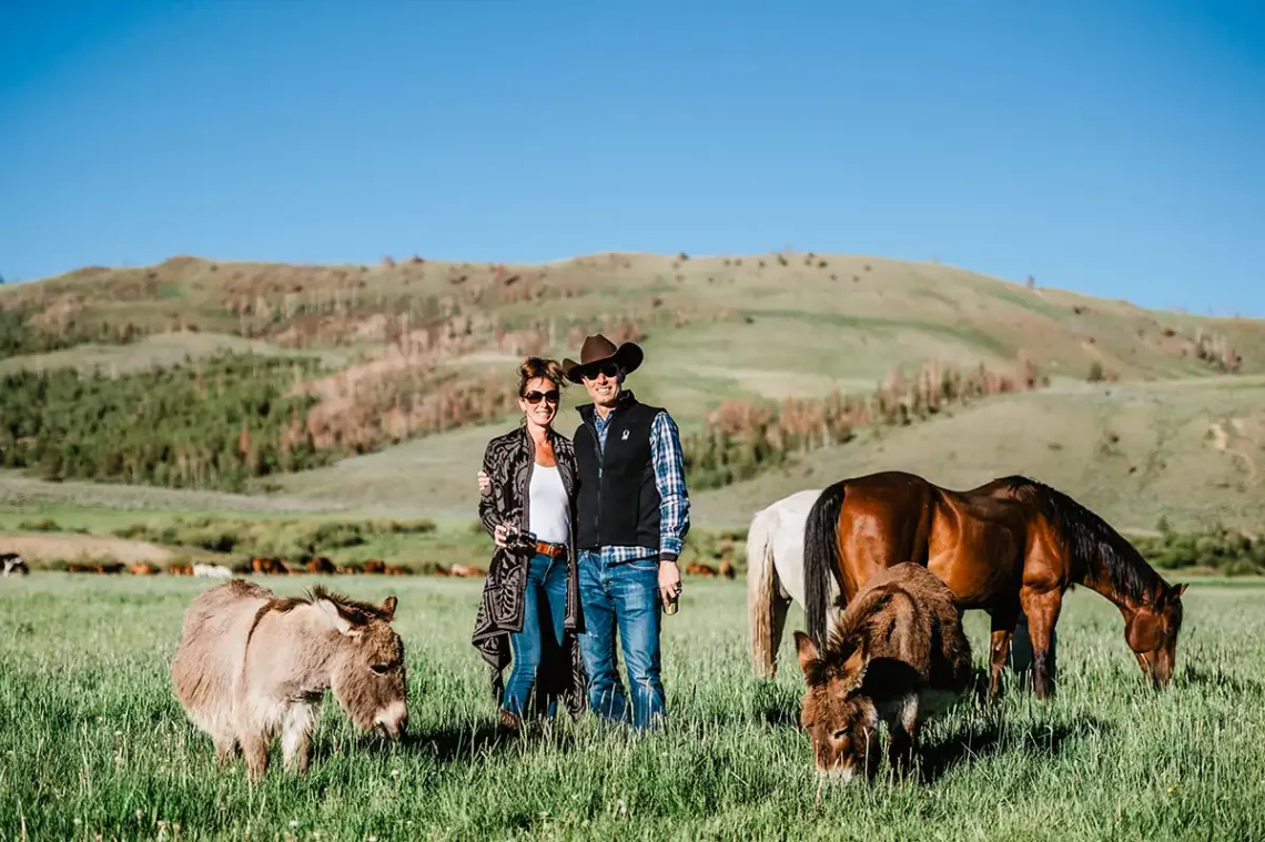 A couple with their arms around each other's waist in the horse pasture with the donkeys at happy hour