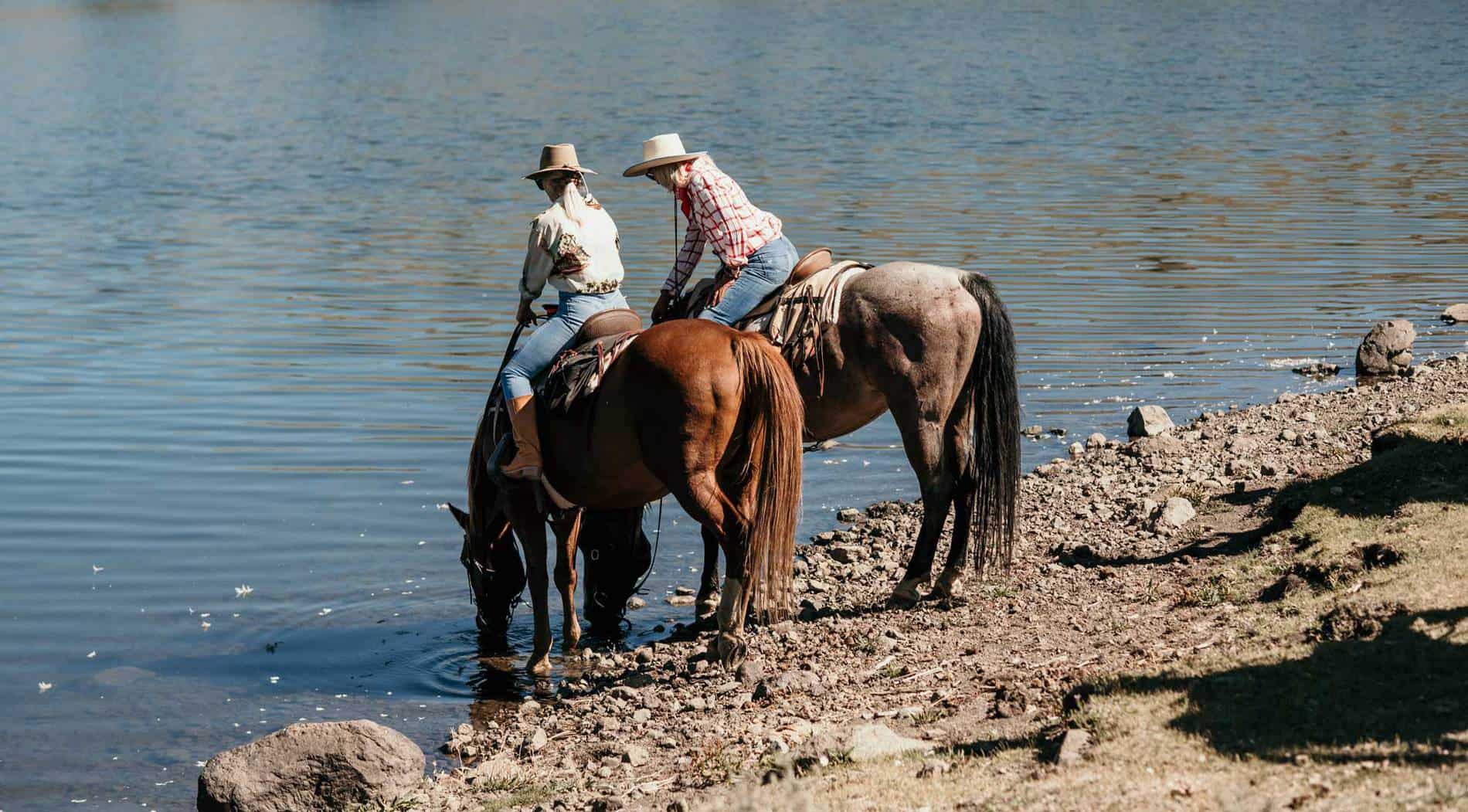 Two women on horses let the horses drink from the lake